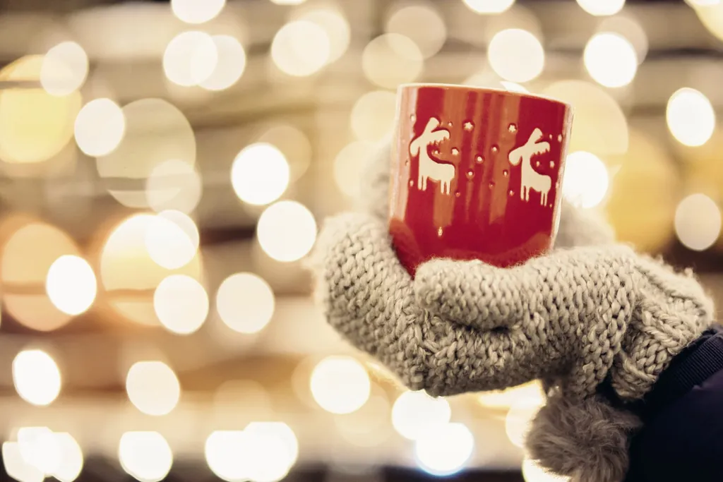 Hand in dickem Wollhandschuh hält eine rote Tasse mit Rentieren, verschwommene Lichter im Hintergrund