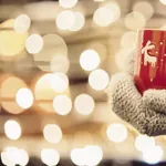 Hand in dickem Wollhandschuh hält eine rote Tasse mit Rentieren, verschwommene Lichter im Hintergrund