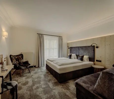 Blick in ein Zimmer des Familienresort Reslwirt in Grautönen mit Holzdetails und großem Doppelbett, gemütlichem Sessel, TV und gepolsteter Sitzbank