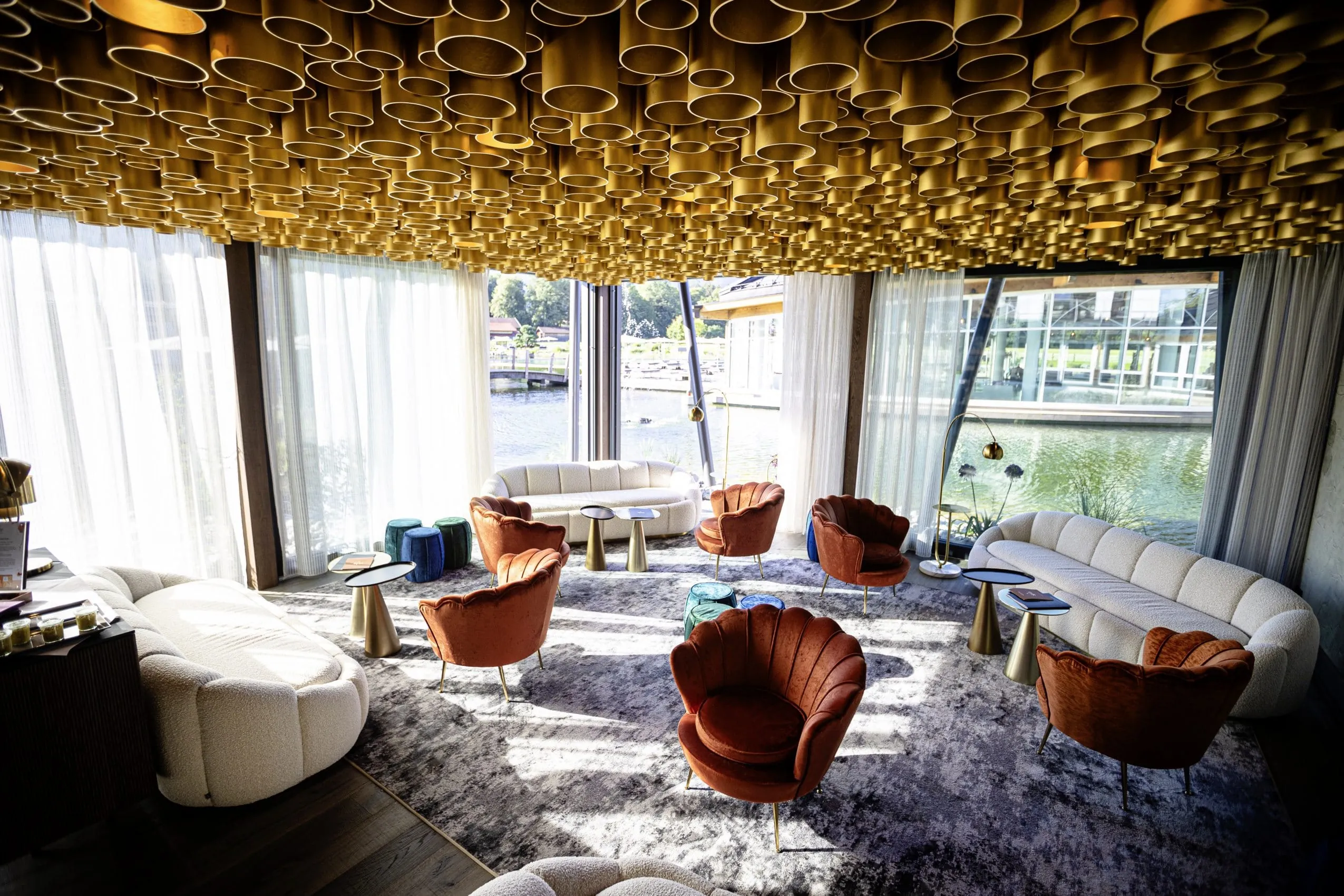 Beigefarbene Couchen um sie herum sind jeweils kleine Tischchen und kupferfarbene Sessel sowie blaue Hocker angeordnet. Die Decke ist ein Designobjekt aus unz&auml;hligen unterschiedlich langen, goldenen R&ouml;hren.