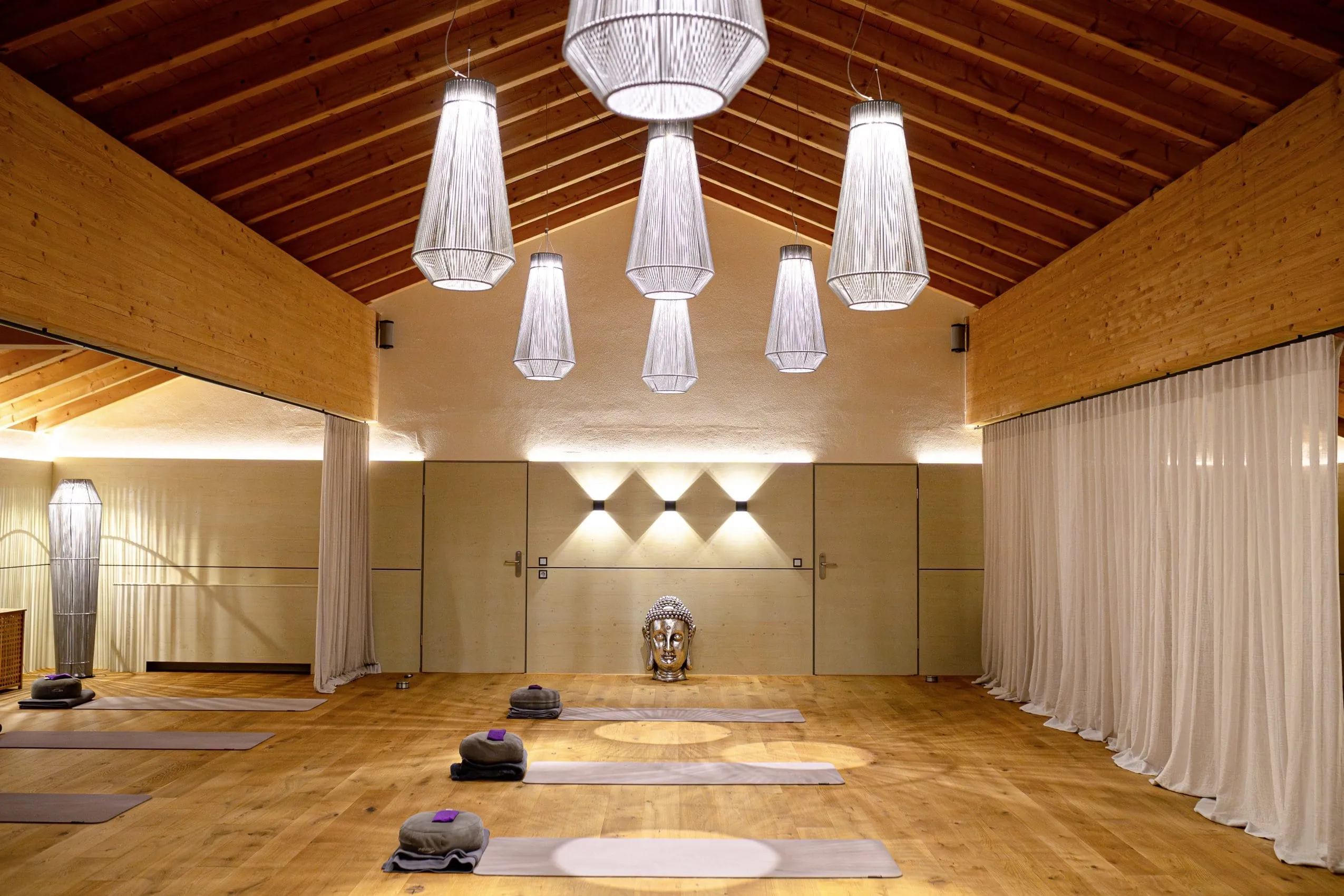 Hoher Saal mit gem&uuml;tlichem Holzboden und gro&szlig;en dekorativen H&auml;ngelampen. Am Boden steht eine Buddha Statue und Yogamatten inklusive Yogakissen und Handtuch liegen parat.
