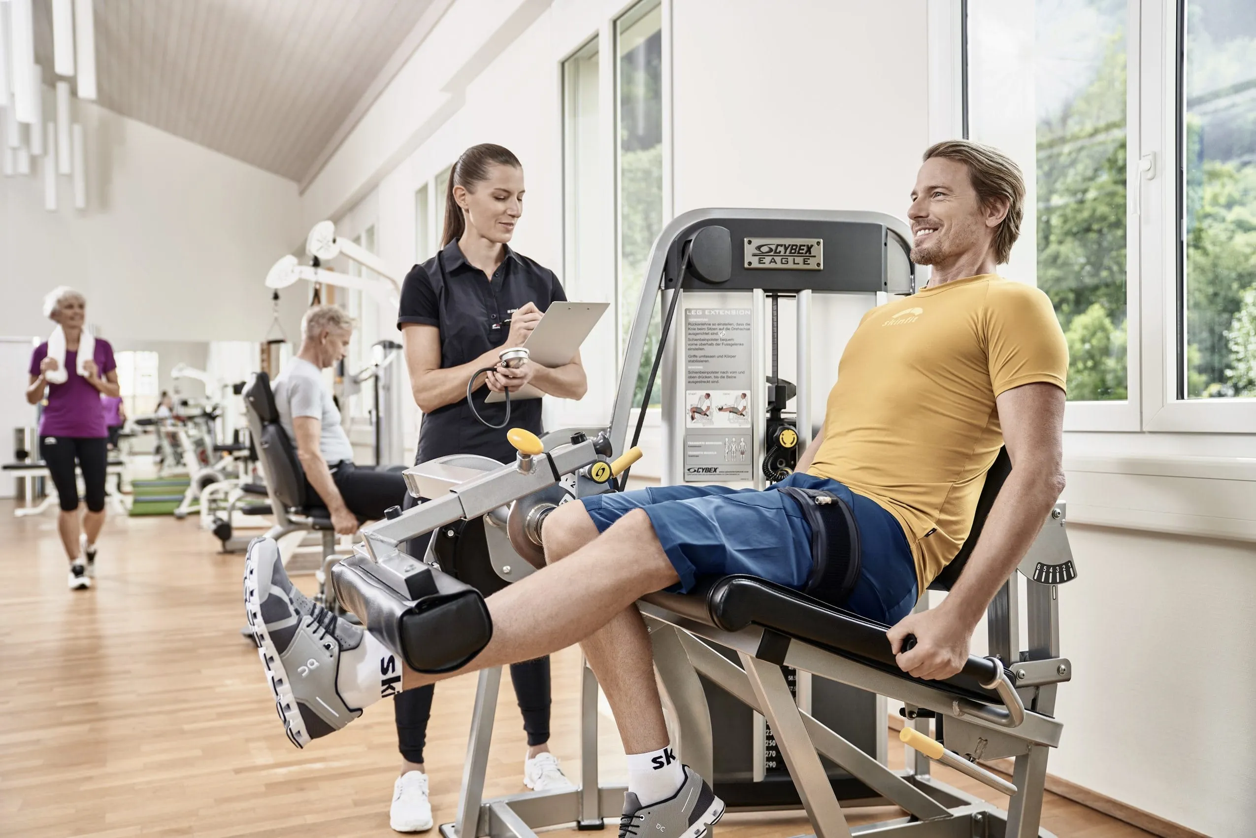 Ein junger, blonder Mann trainiert an einem Fitnessger&auml;t seine Beine. Daneben steht eine Personaltrainerin mit Klemmbrett und Stoppuhr in der Hand, die ihn betreut.