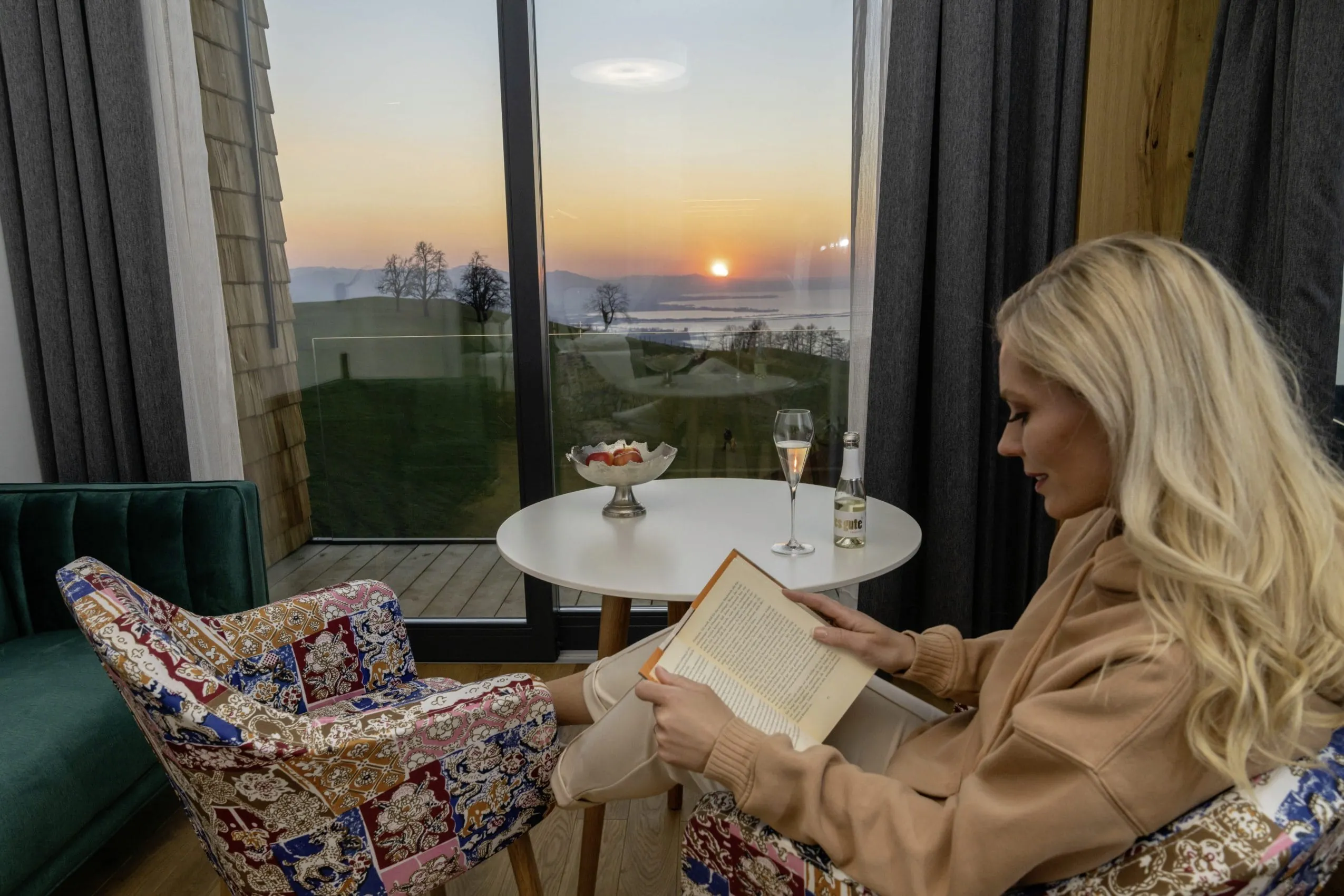 Eine junge Frau mit blonden, langen Haaren sitzt gem&uuml;tlich in einem bunten Sessel und liest ein Buch vor dem gro&szlig;en Terrassenfenster. Drau&szlig;en f&auml;rbt der Sonnenuntergang den Himmel in bunten Farben.