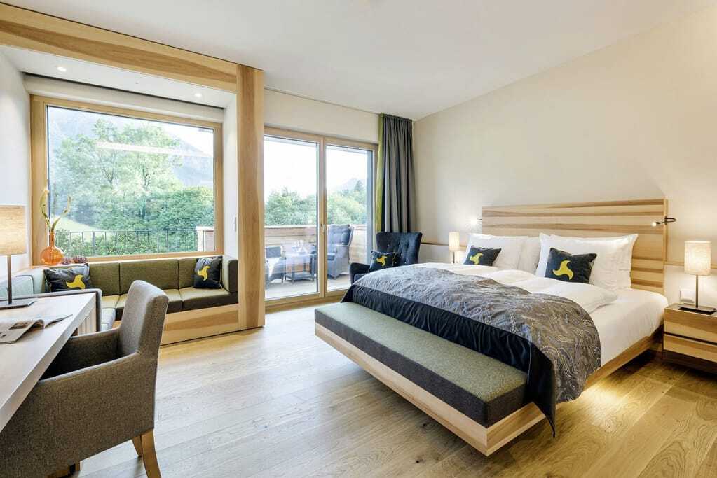 Hotelzimmer mit hellem Paketboden großem Panoramafenster zum Balkon und gemütlicher Sitznische mit grünen Polstern.