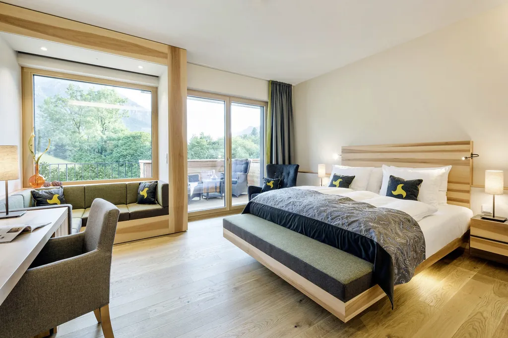 Hotelzimmer mit hellem Paketboden gro&szlig;em Panoramafenster zum Balkon und gem&uuml;tlicher Sitznische mit gr&uuml;nen Polstern.