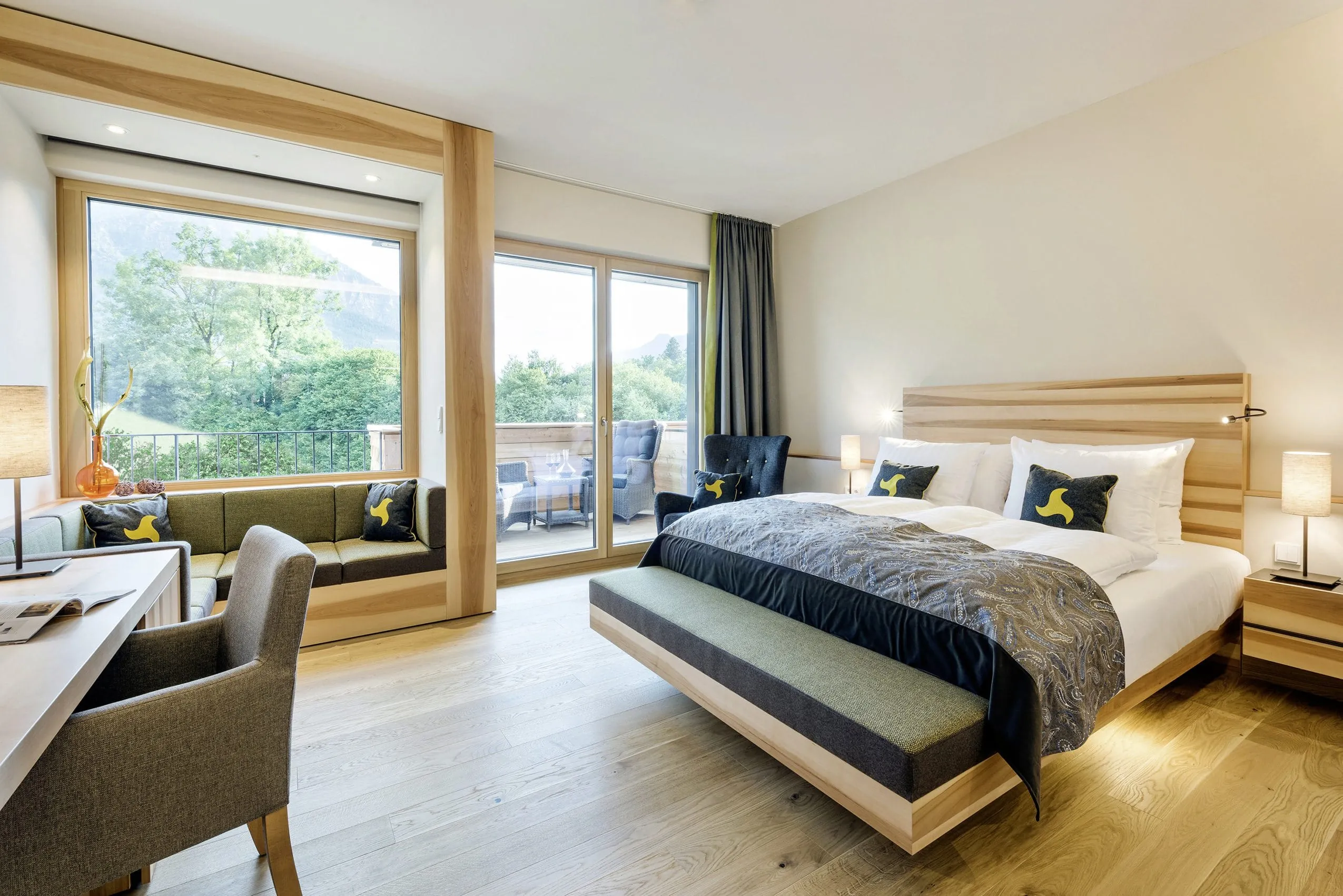 Hotelzimmer mit hellem Paketboden gro&szlig;em Panoramafenster zum Balkon und gem&uuml;tlicher Sitznische mit gr&uuml;nen Polstern.