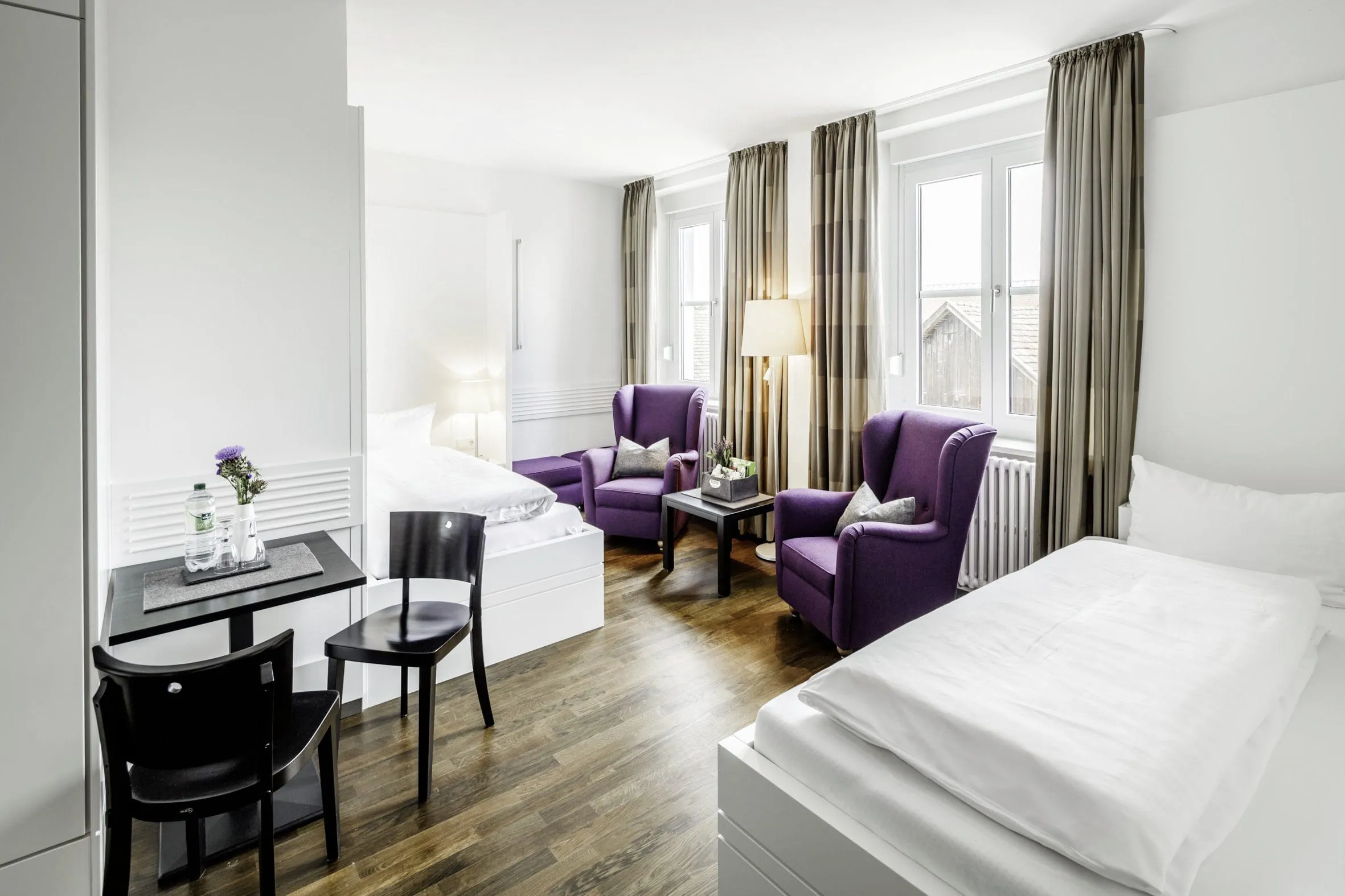 Sehr clean gehaltenes Zimmer. Nur die lilanen Sessel und beigen Vorh&auml;nge setzen farbliche Akzente im sonst wei&szlig; gehaltenen Raum.