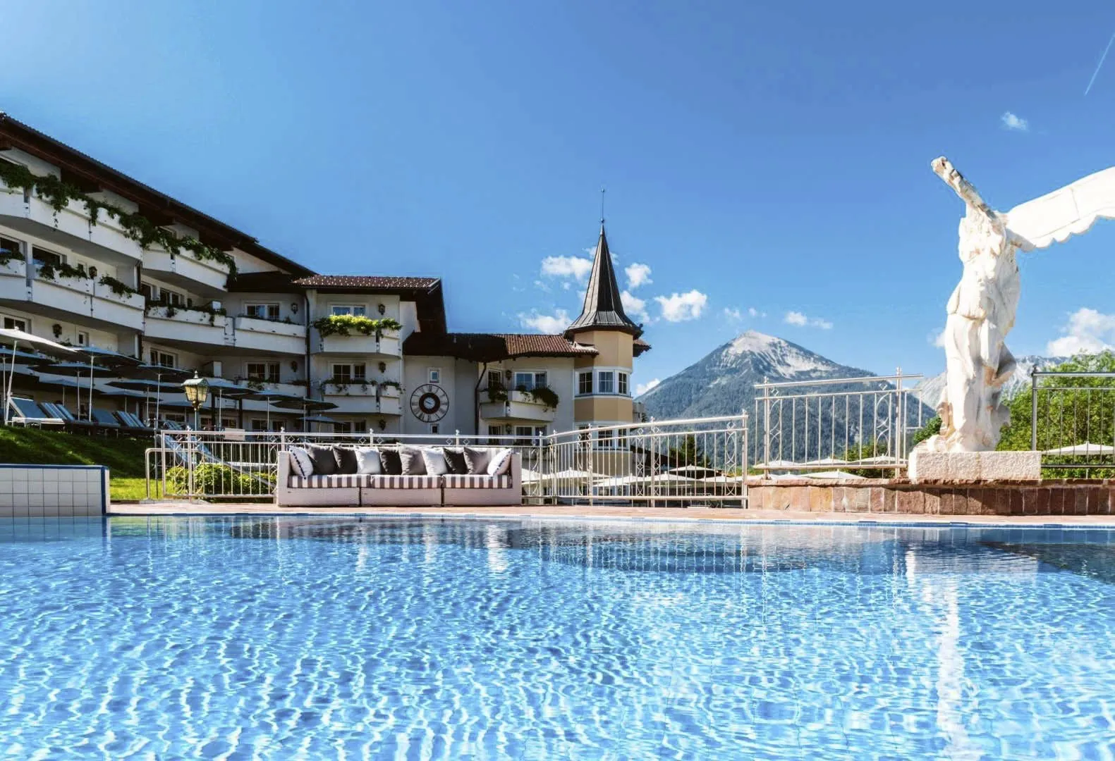 Das blaue Wasser des Pools glitzert in der Sonne. Im Hintergrund ist das Hotelgeb&auml;ude mit zahlreichen Balkonen und die Berge zu sehen.