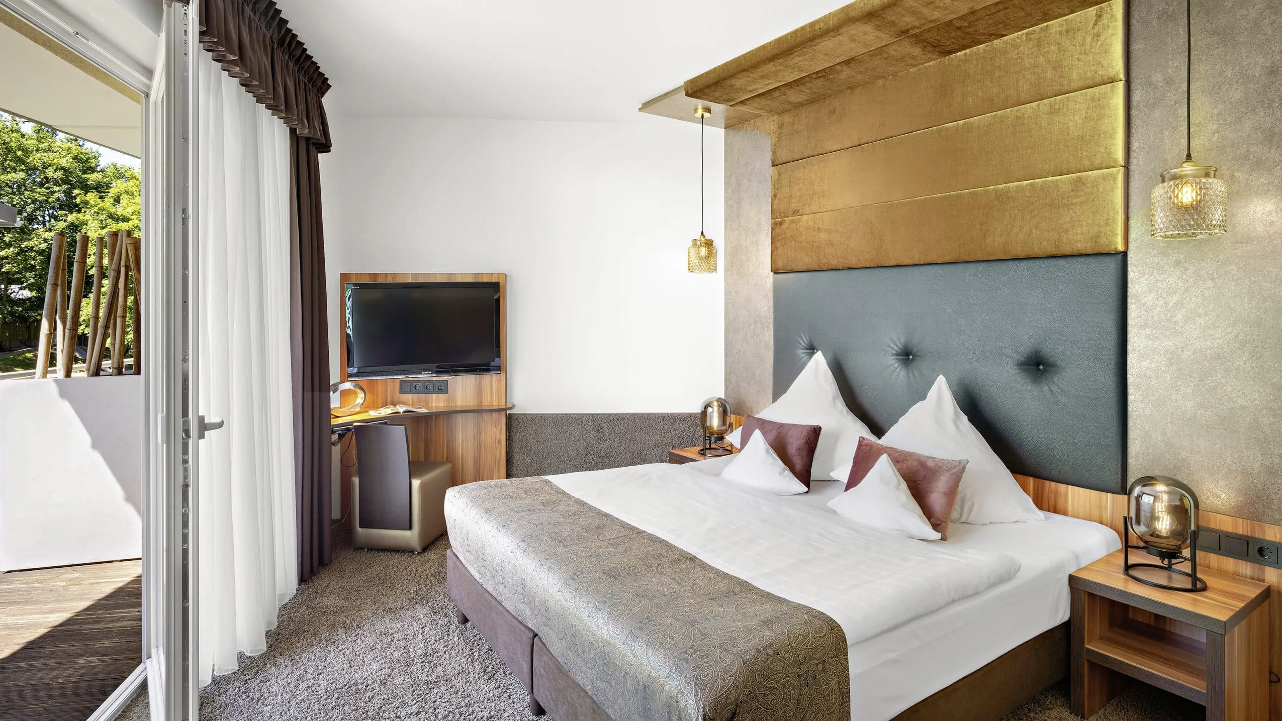 Gro&szlig;es Boxspringbett mit braunen Polstern, Wandverkleidung in Messing-Metaloptik, hellbrauner Hochflorteppich und gro&szlig;er Balkont&uuml;re.