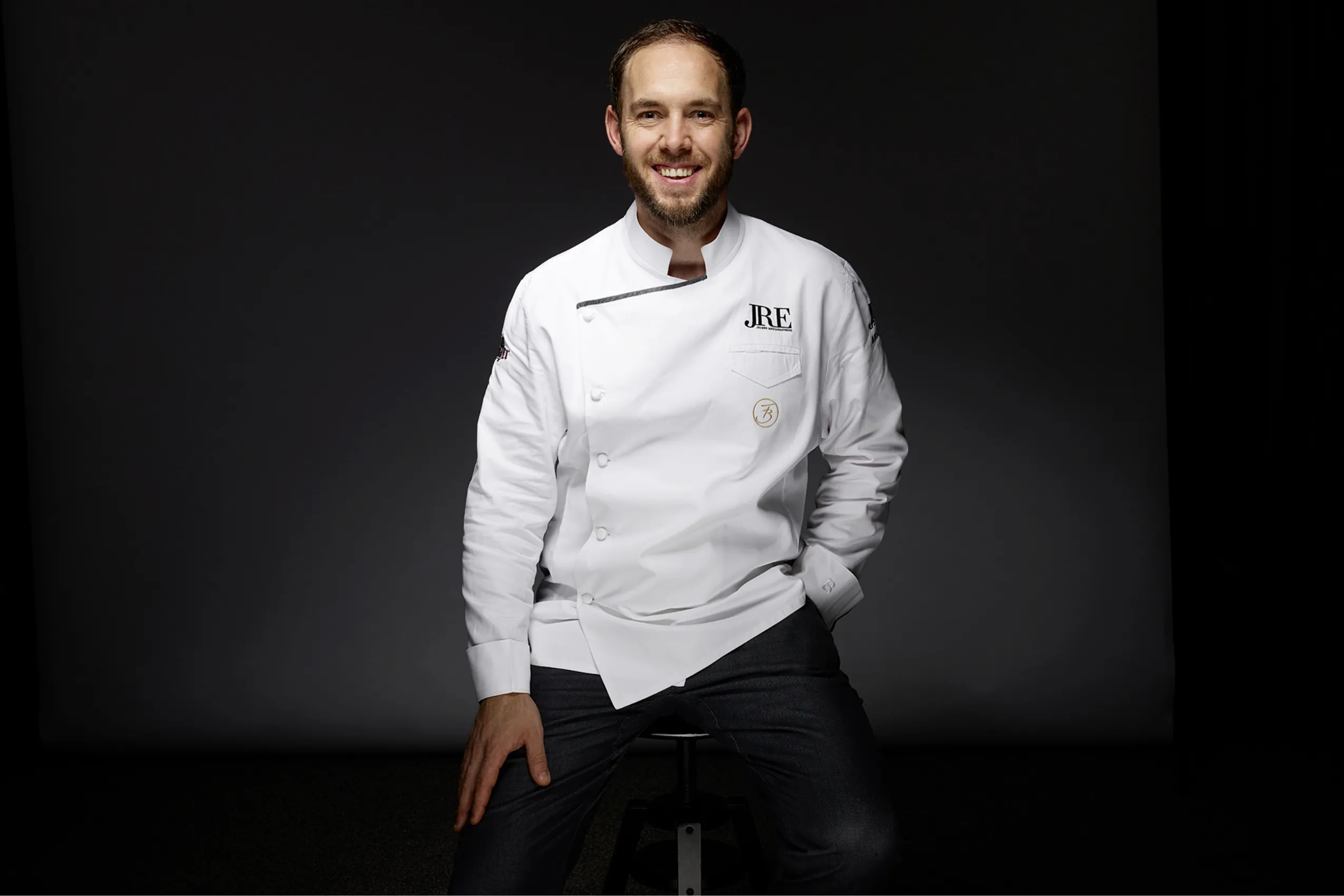 Portr&auml;t von K&uuml;chenchef Franz Berlin l&auml;ssig auf einem Hocker vor schwarzem Hintergrund