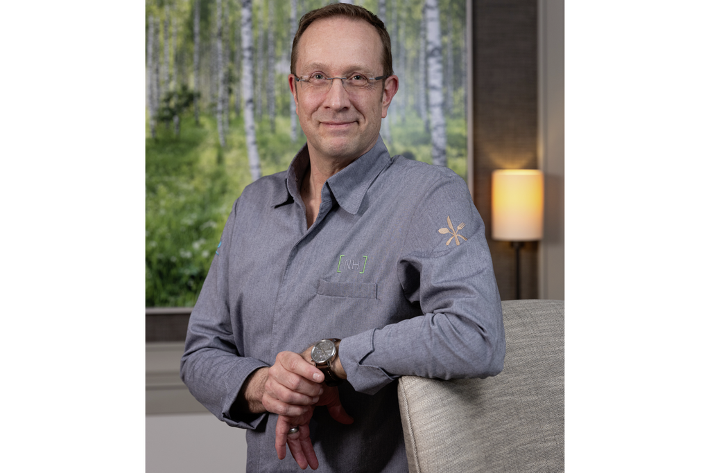 Sternekoch Nils Henkel lächelnd im Halbfigurenporträt mit grauem Kochhemd vor Gemälde mit Birken im Sternerestaurant Tipkens by Nils Henkel