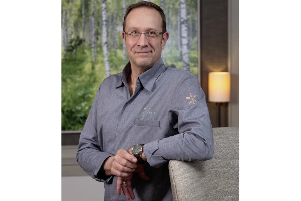 Sternekoch Nils Henkel l&auml;chelnd im Halbfigurenportr&auml;t mit grauem Kochhemd vor Gem&auml;lde mit Birken im Sternerestaurant Tipkens by Nils Henkel