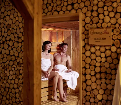 Paar sitzt mit weißen Handtüchern in der Sauna des Vitalhotel Sonneck mit Holzscheiten vor dem Eingang