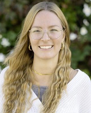 Autorenporträt Lena Schmidberger mit runder Brille, langen, welligen Haaren und weißer Strickjacke