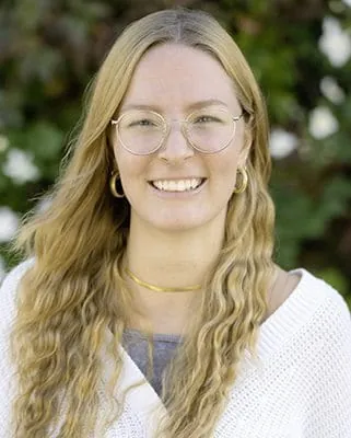 Autorenporträt Lena Schmidberger mit runder Brille, langen, welligen Haaren und weißer Strickjacke