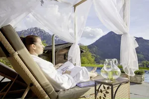 Frau in weißem Bademantel liegt mit geschlossenen Augen auf einem Daybed am Teich