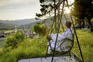 Frau in weißem Bademantel sitzt auf Hollywoodschaukel und schaut auf das Dorf