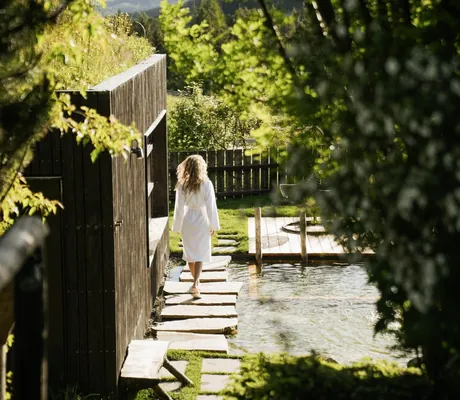 blonde Frau mit weißem Bademantel läuft auf Steinen am Teich des Hotel Pfösl entlang