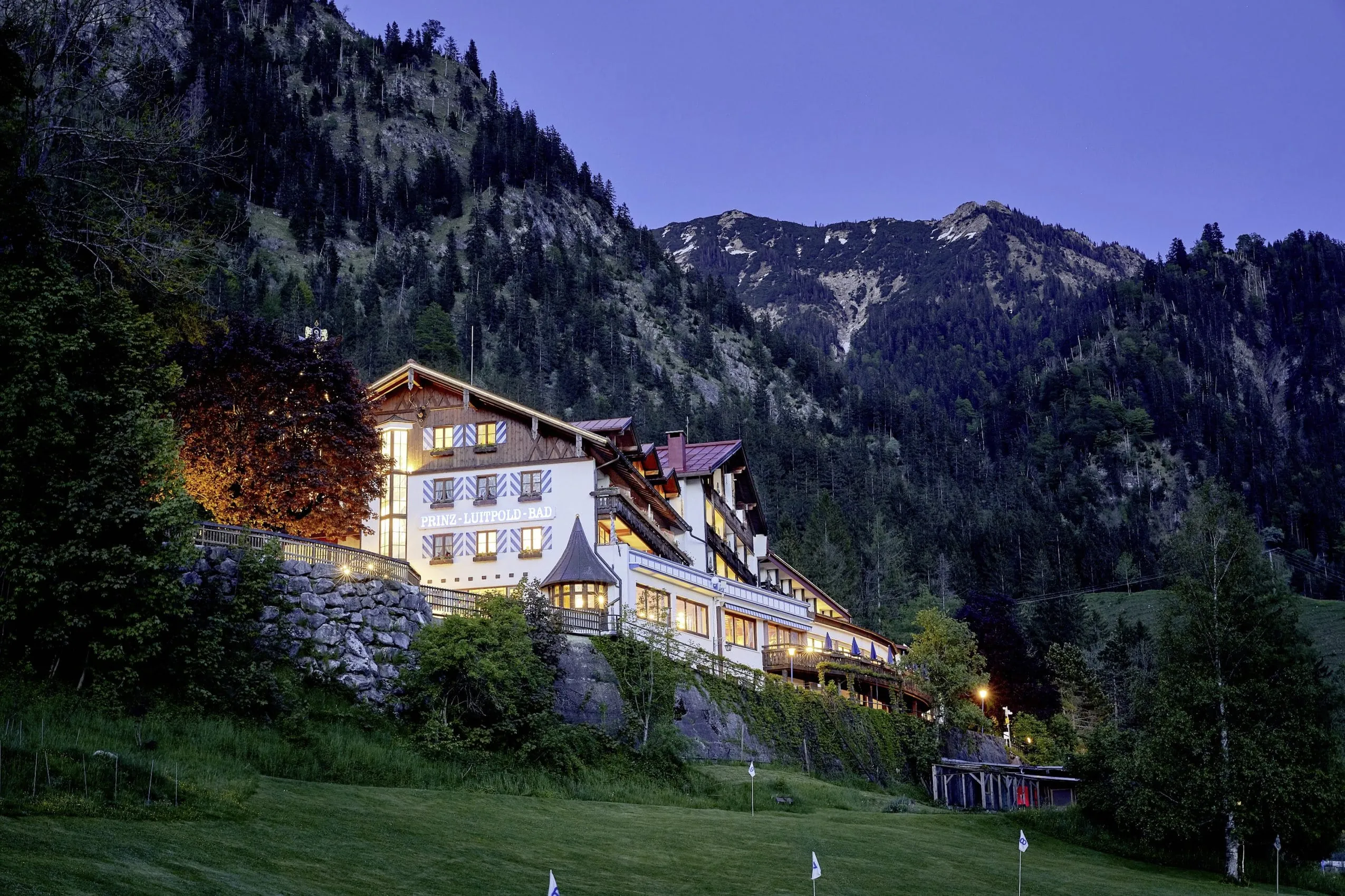 Au&szlig;enaufnahme des Hotels am Abend wie es eingebettet in die Allg&auml;uer Alpen auf einer Anh&ouml;he thront. Aus den Fenstern strahlt helles Licht.