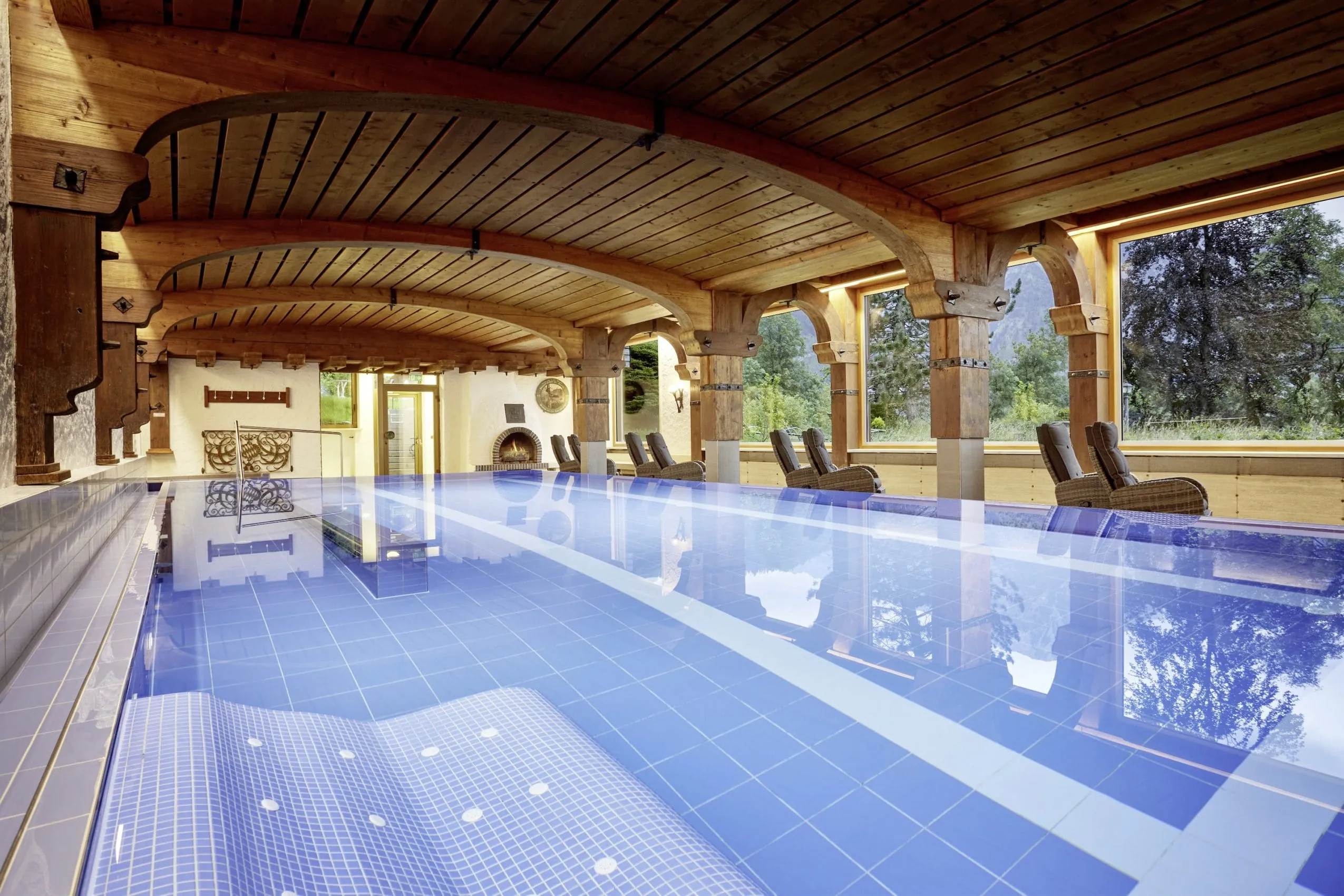 Indoorpool mit blauen Fliesen, Liegefl&auml;che, holzverkleideter Decke und S&auml;ulen sowie gem&uuml;tlichen Sesseln zum Entspannen
