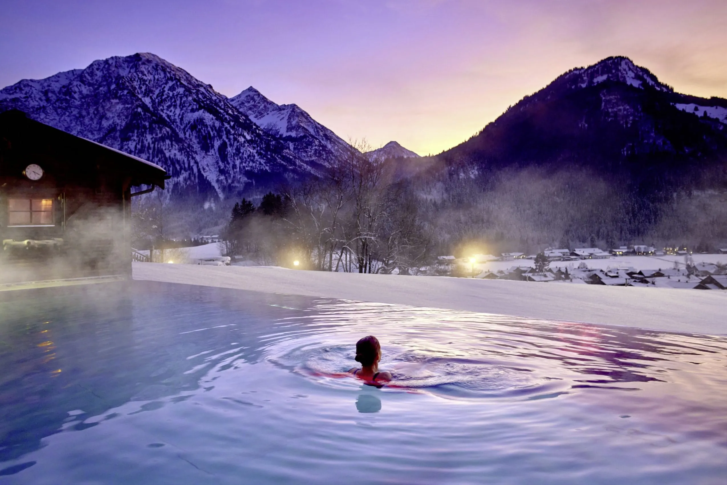 Outdoorpool im Winter. Nebelschwaden bilden sich &uuml;ber dem Pool wegen des Temperaturunterschiedes vom warmen Wasser zur kalten Luft. Im Hintergrund sind schneebedeckte Berge zu sehen.