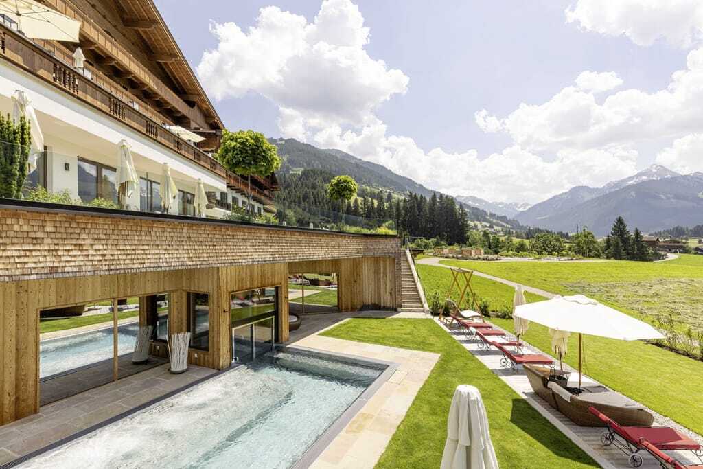 Die holzverkleidete Außenfassade des Tiroler Wellnesshotels Alpbacherhofs mit direkt davorliegendem Außenpool und Liegebereich mit Blick auf die Berge.