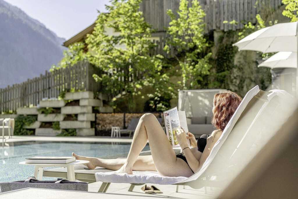 Eine junge Frau entspannt auf einer Liege am Außenpool des Designhotels Bergland in Tirol und liest ein Magazin.