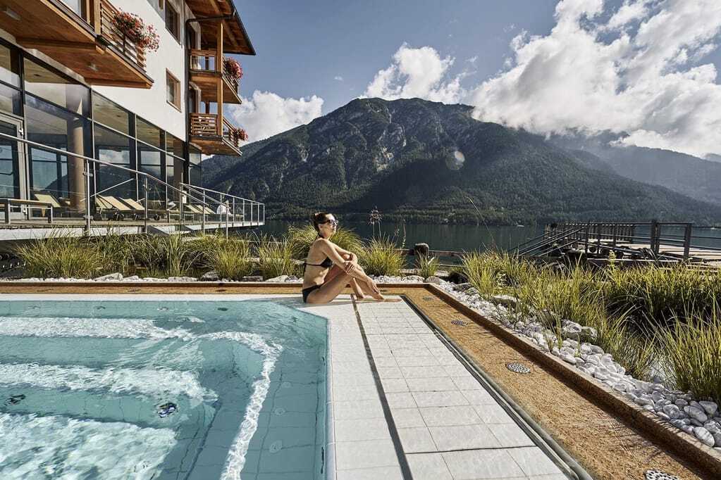 Eine Frau genießt am Beckenrand des Außenpools die Sonne. Direkt daneben liegt das Hotelgebäude des Wellnesshotels Fürstenhaus sowie der Achensee.