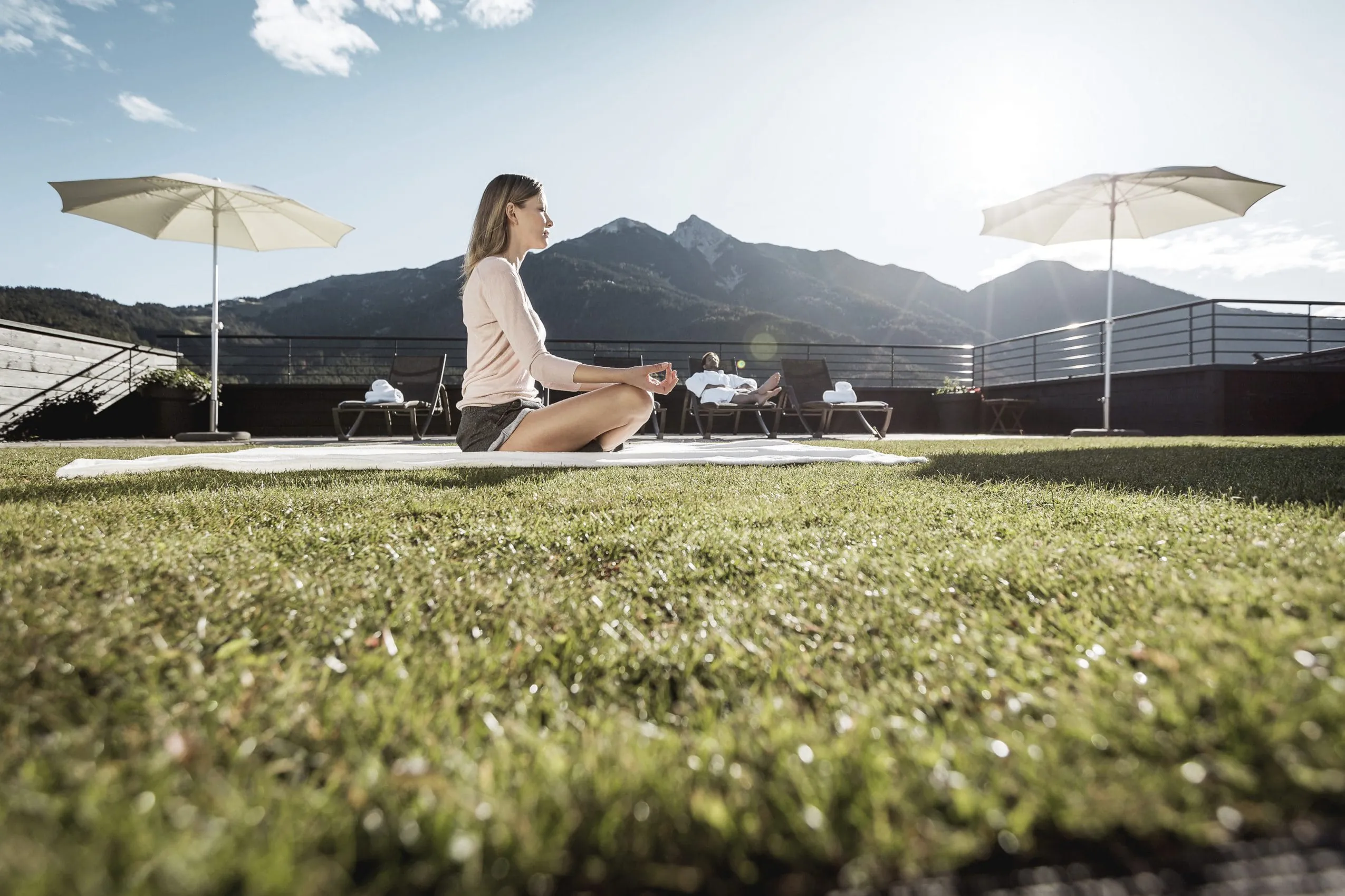 Die begr&uuml;nte Dachterrasse des Post Seefelds l&auml;dt mit Liegen und Sonnenschirmen zum Entspannen ein. Eine Frau meditiert auf einer Decke. Ein Mann entspannt im Bademantel auf einer Liege im Hintergrund