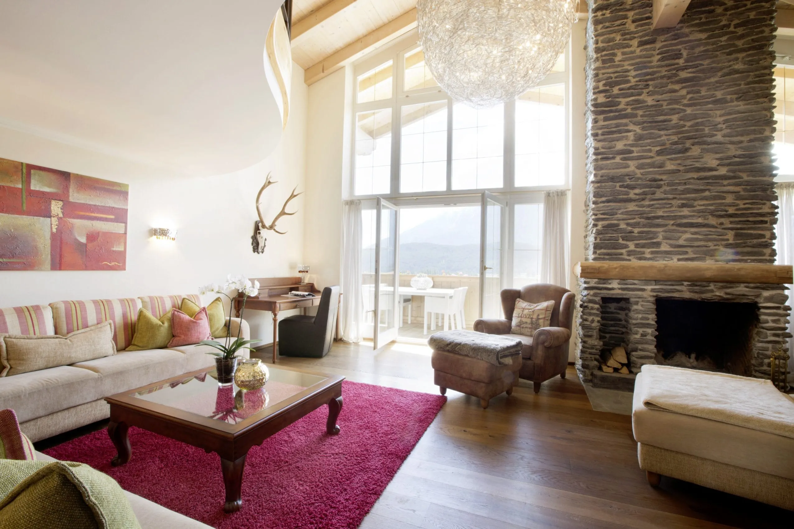 Wohnbereich der Royal Garden Suite mit hohen Fenstern und Kamin aus grobem Naturstein. Davor befindet sich ein helles Sofa und ein Ohrensessel f&uuml;r gem&uuml;tliche Stunden.