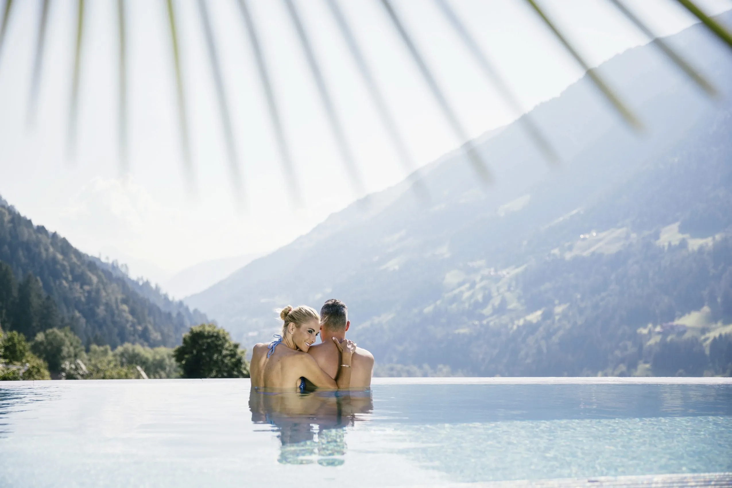 Infinity-Outdoorpool in dem sich ein P&auml;rchen z&auml;rtlich aneinander schmiegt und die umliegenden Berge genie&szlig;t.