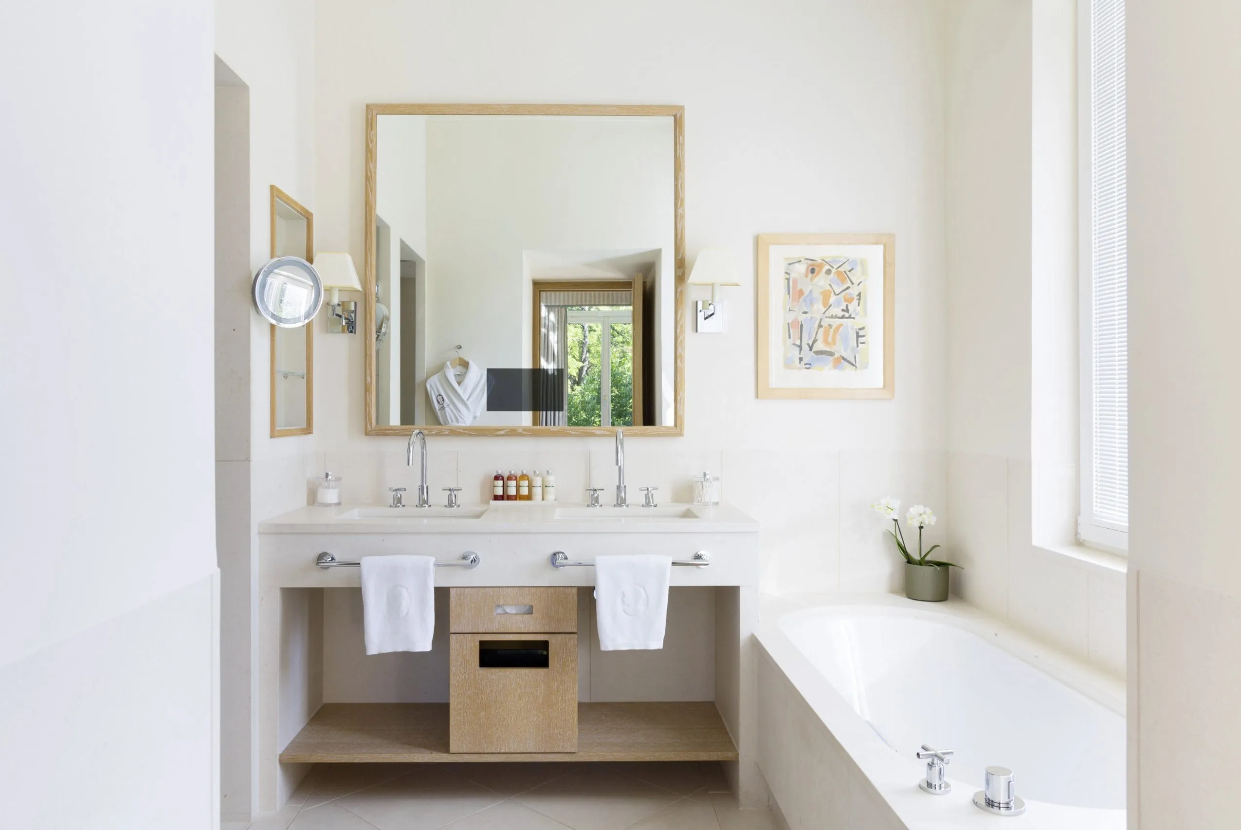 Helles, lichtdurchflutetes Badezimmer mit hellbeigen Fliesen, Doppelwaschbecken, Badewanne und gro&szlig;em Spiegel mit Holzrand.