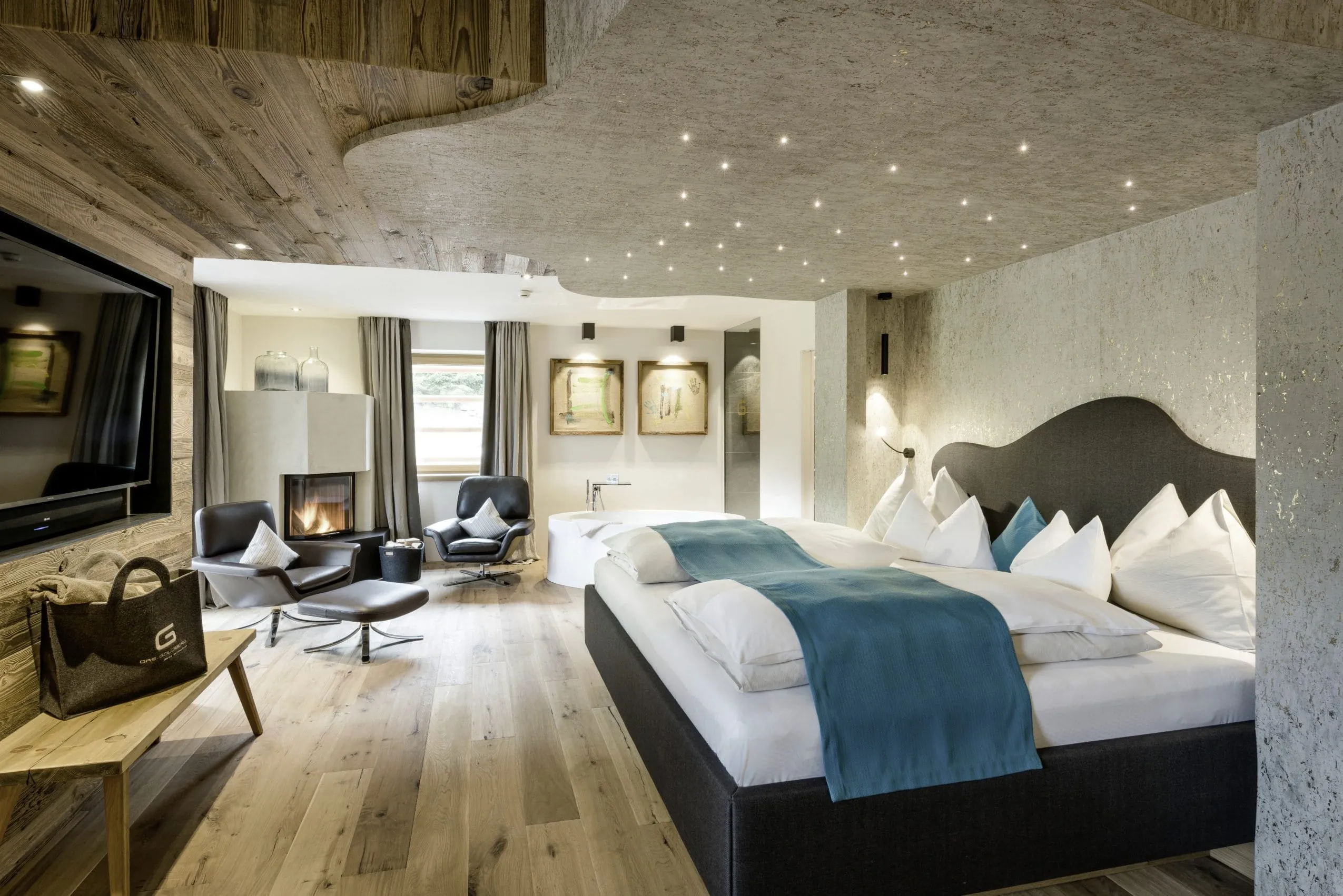 Schlafzimmer mit einer Betonoptik- und einer Holzwand, gro&szlig;em schwarzen Kingsizebett. Eine freistehende Badewanne und ein Kamin mit zwei gem&uuml;tlichen schwarzen Ledernsesseln davor befinden sich am anderen Ende des Raums.