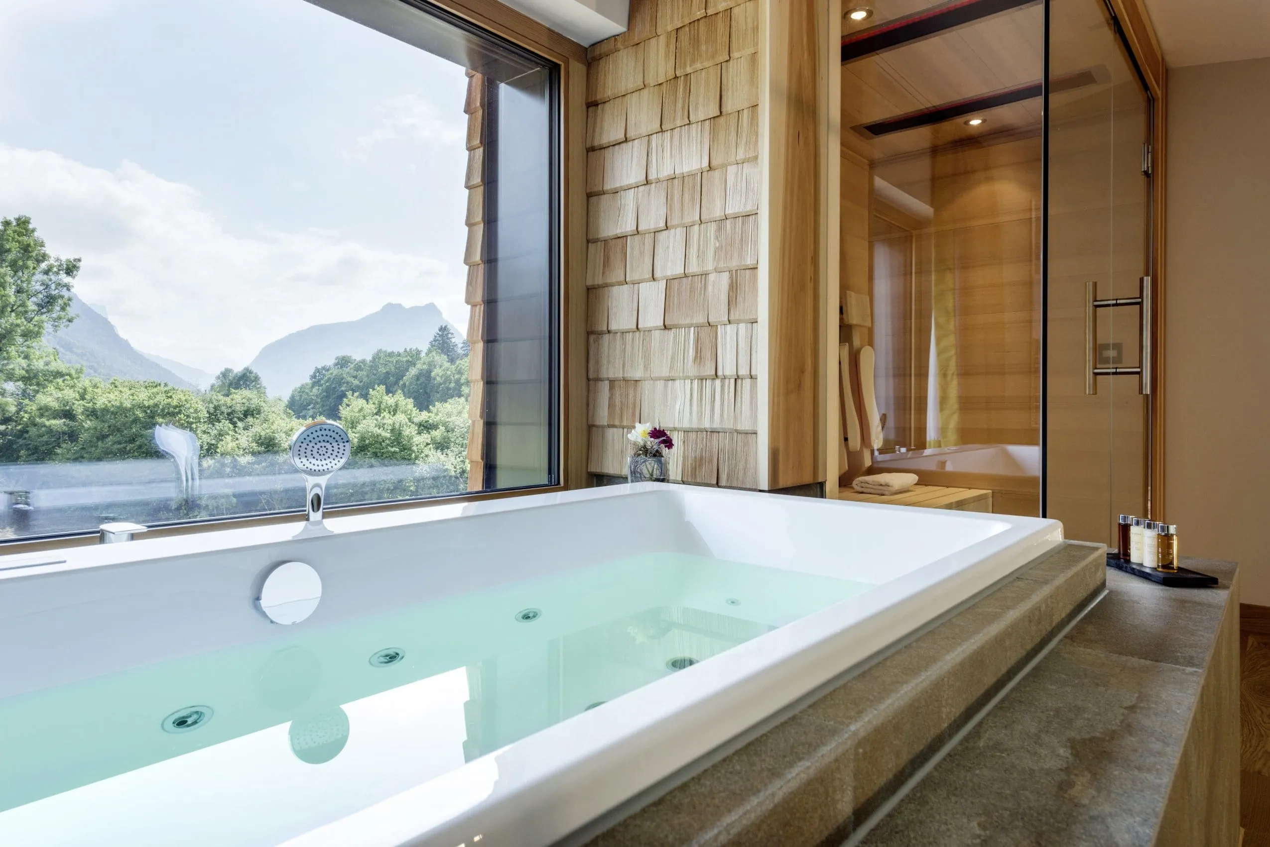 Whirlpoolbadewann in Natursteinfliesen eingelassen neben gro&szlig;em Panoramafenster mit Blick auf die Berge.