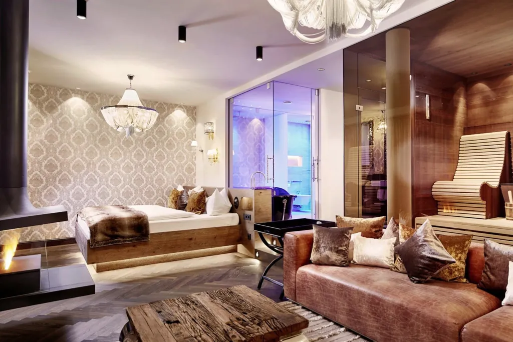 Privat Wellness Suite mit großzügigem Badezimmer und Sauna abgetrennt durch eine Glaswand vom Wohnbereich mit Schlafzimmer, großem Sofa und Kamin.