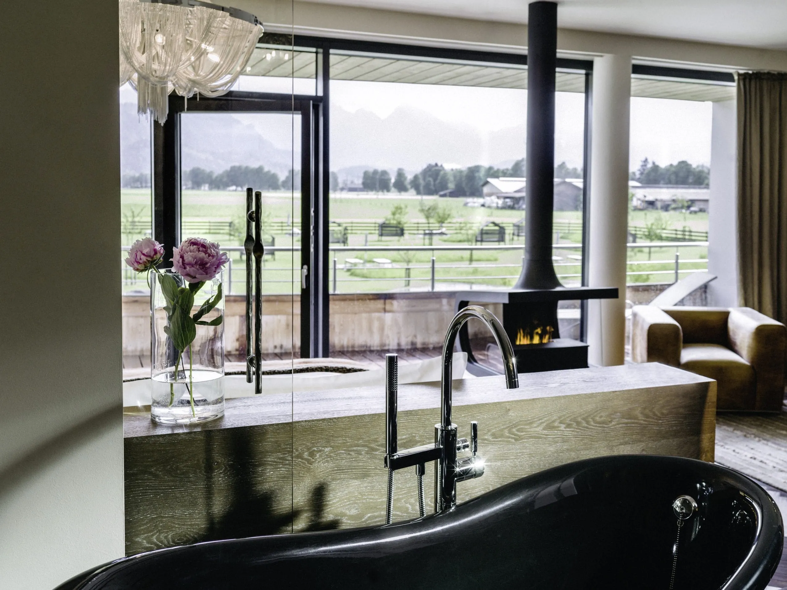 Freistehende schwarze Badewanne hinter einer halbhohen Trennwand. Dahinter Wohnraum mit Blick auf die Terrasse und den Kamin.
