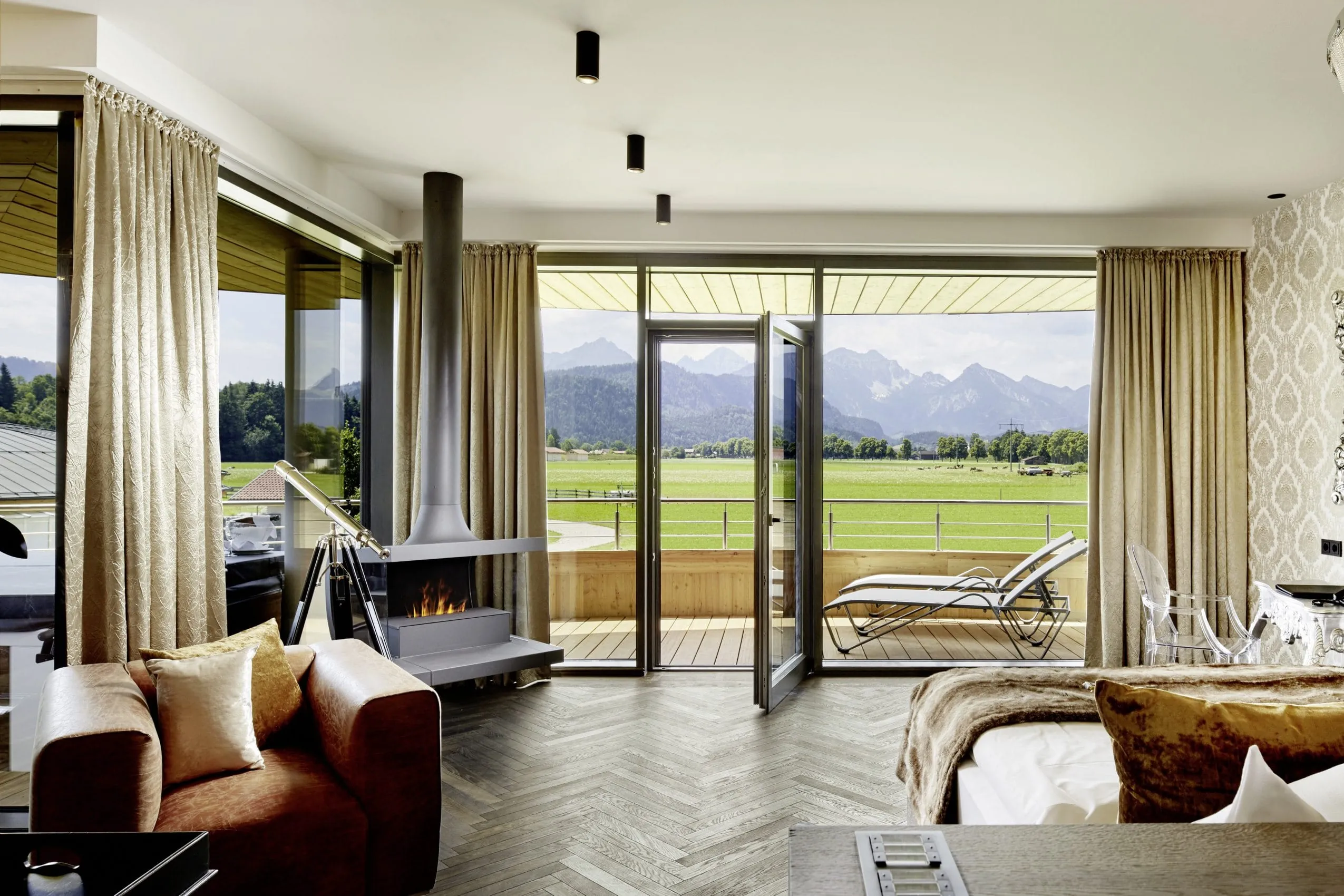 Gro&szlig;es Terrassenfenster mit ge&ouml;ffneter T&uuml;r, die zauberhafte Blicke auf die Berge zul&auml;sst. Wohnbereich mit braunem Ledersessel und gem&uuml;tlichem Kamin.