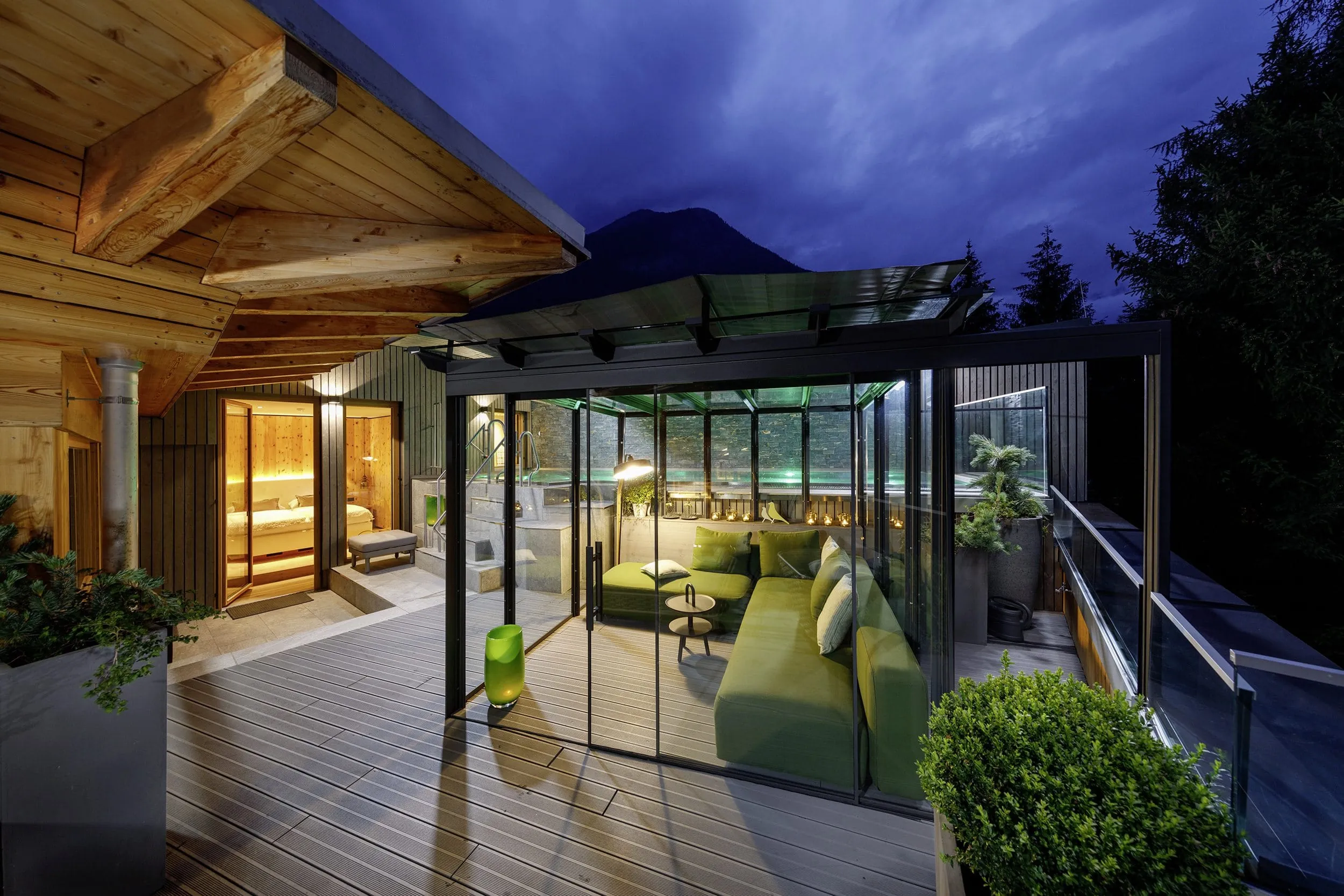 Terrasse mit gr&uuml;nem Lounge-Sofa im Wintergarten. Dahinter ein Pool und Bergpanorama in der Abendd&auml;mmerung.