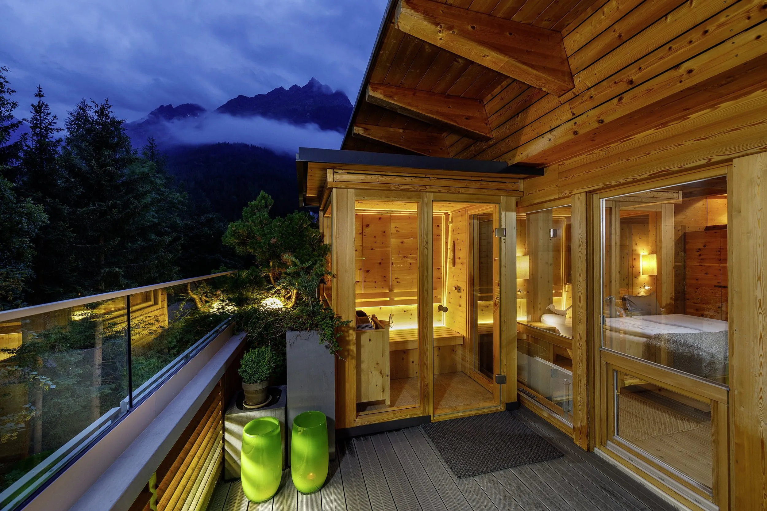Balkon mit privater, kleiner Sauna in der Abendd&auml;mmerung. Im Hintergrund spitzen hohe Berggipfel durch weit unten h&auml;ngende Wolken.