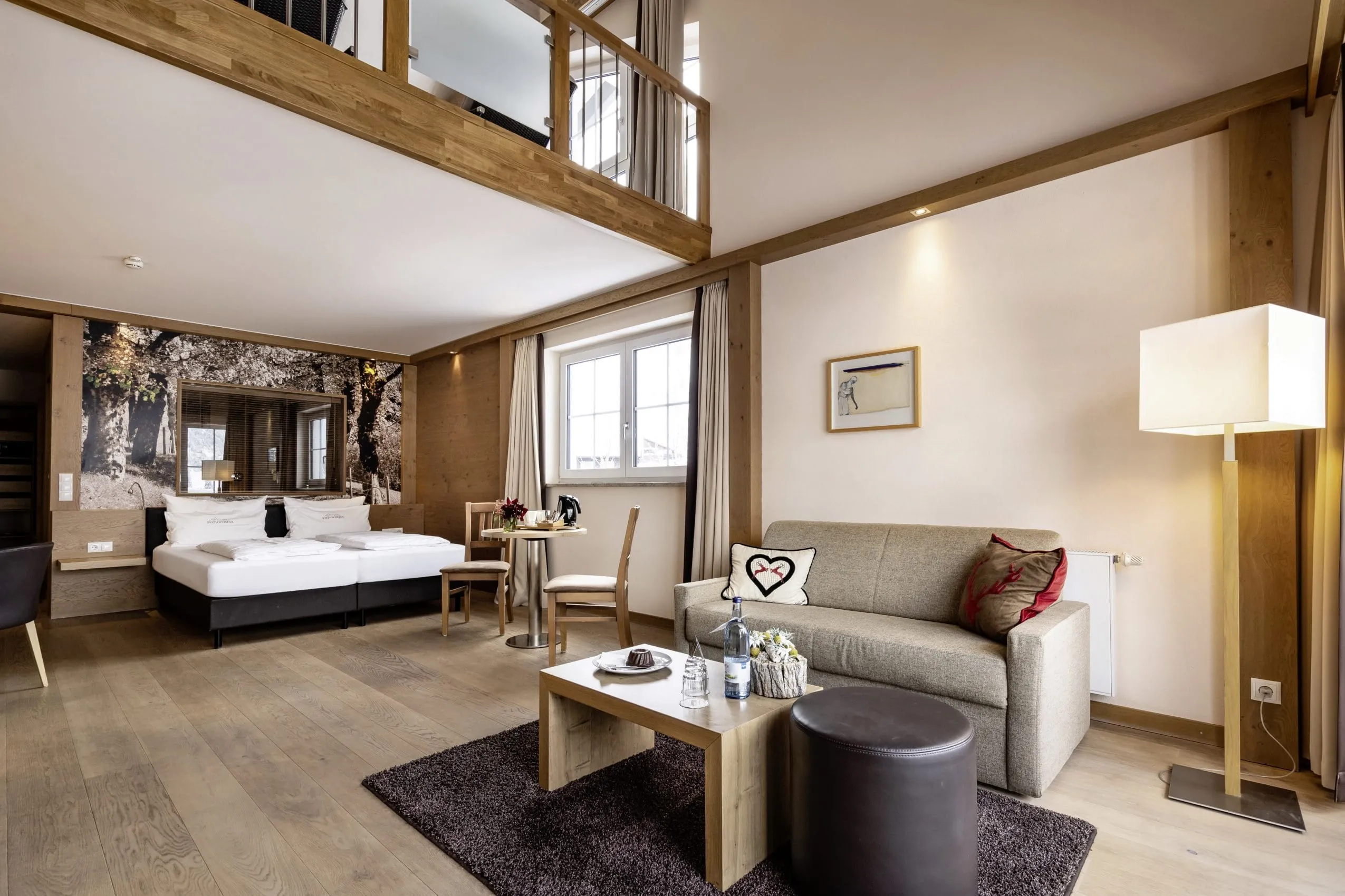 Gro&szlig;es Doppelbett mit wei&szlig;er Bettw&auml;sche. Dar&uuml;ber liegt eine Galerie. Im Vordergrund Wohnbereich mit kleinem grauen Sofa und Couchtisch.