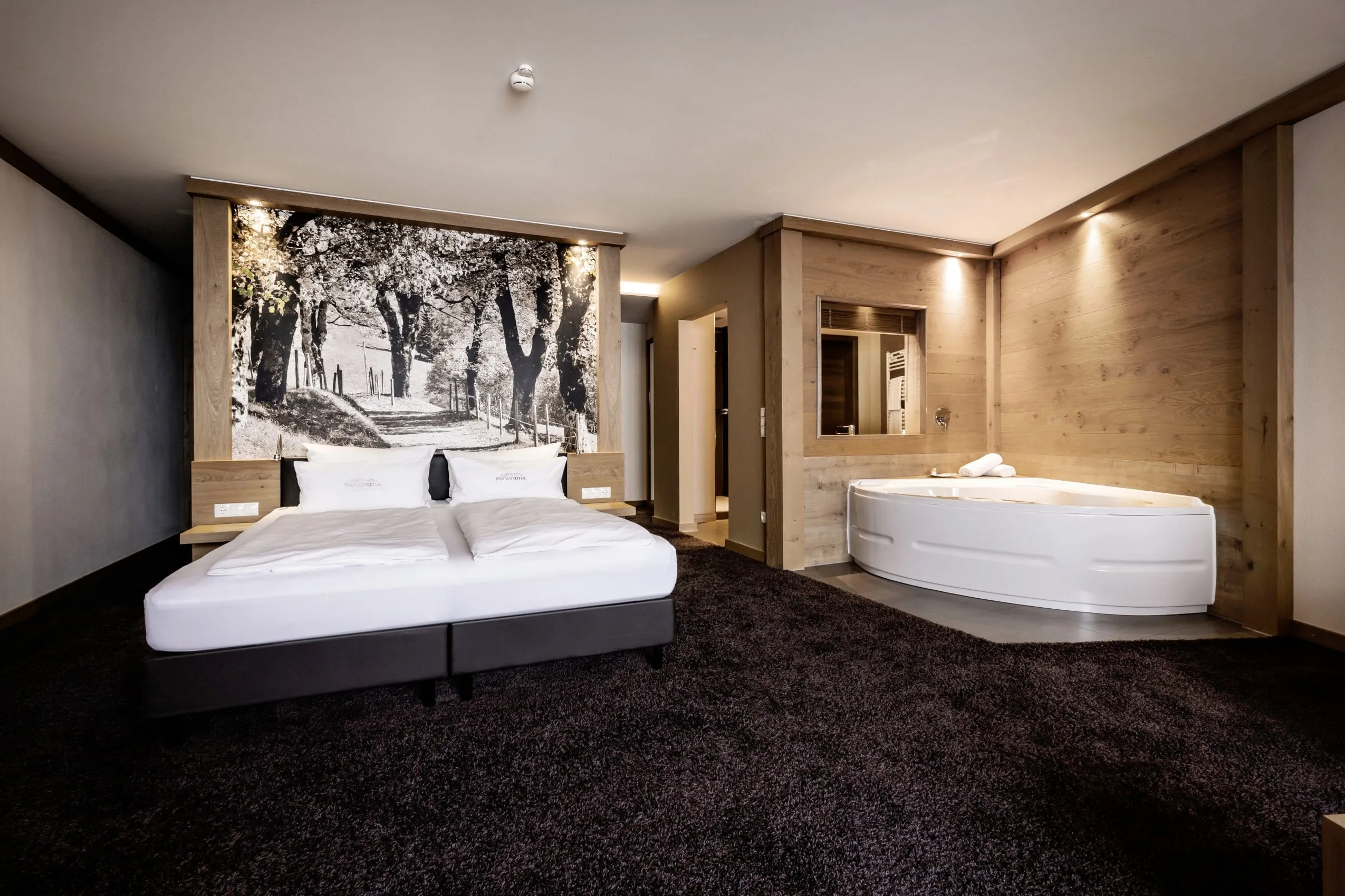 Schlafzimmer mit Holzw&auml;nden und dunklem Hochflor-Teppich. Gro&szlig;es Kingsizebett mit wei&szlig;er Bettw&auml;sche und Eckwhirlpoolbadewanne in der Ecke daneben.