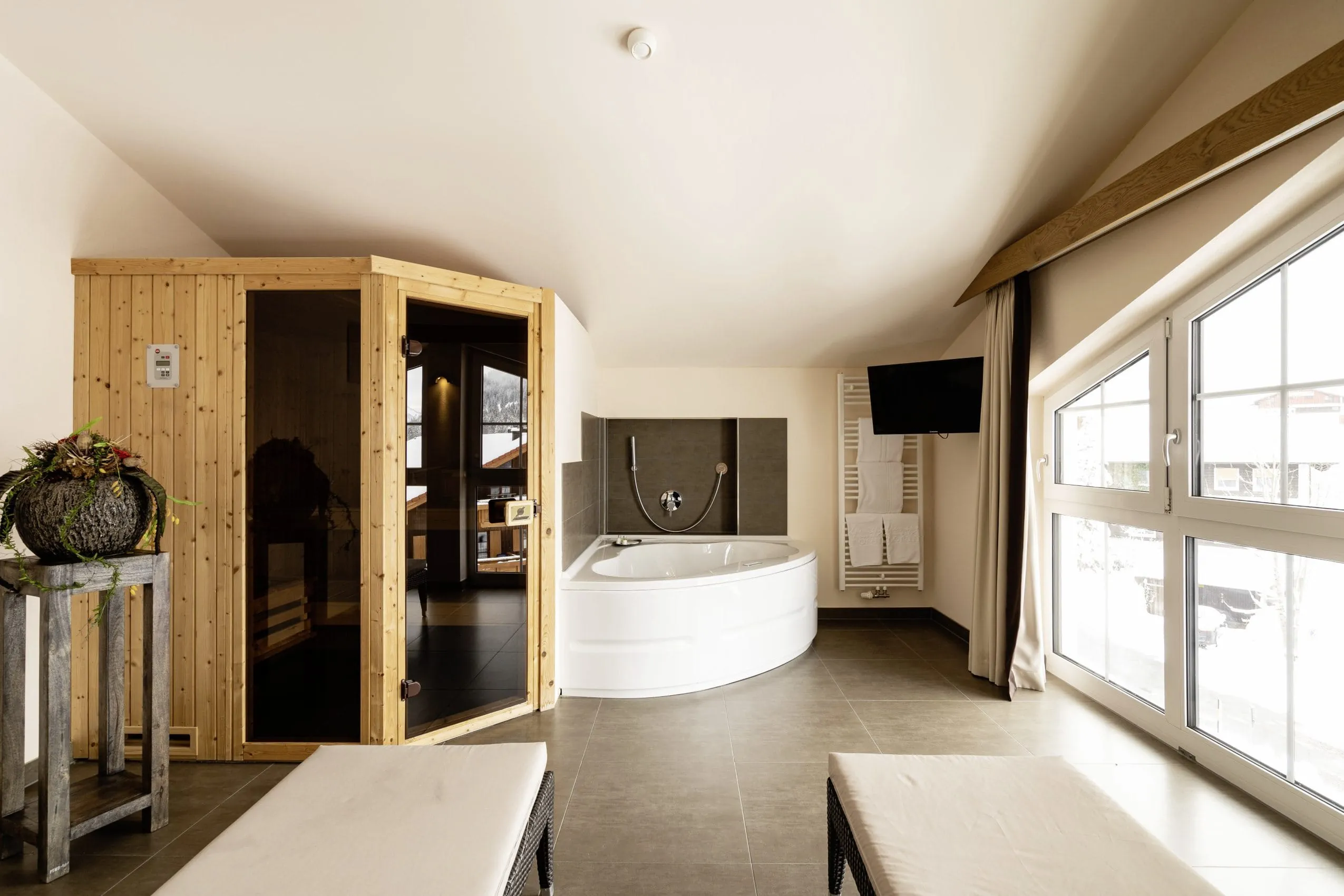 Whirlpoolbadewanne und Sauna in einem Raum mit gro&szlig;en dunklen Fliesen, Panoramafenster und Ruheliegen.