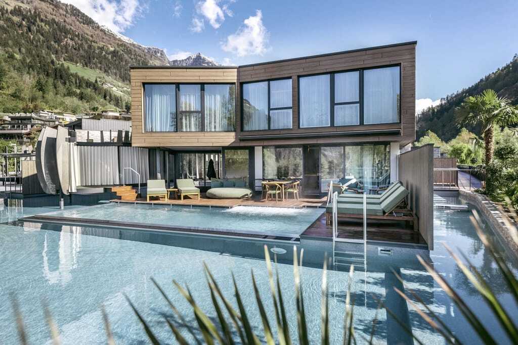 Modernes Gebäude mit Flachdach und kompletter Fensterfront. Davor liegt die Terrasse mit Ruheliegen und davor liegendem Pool.
