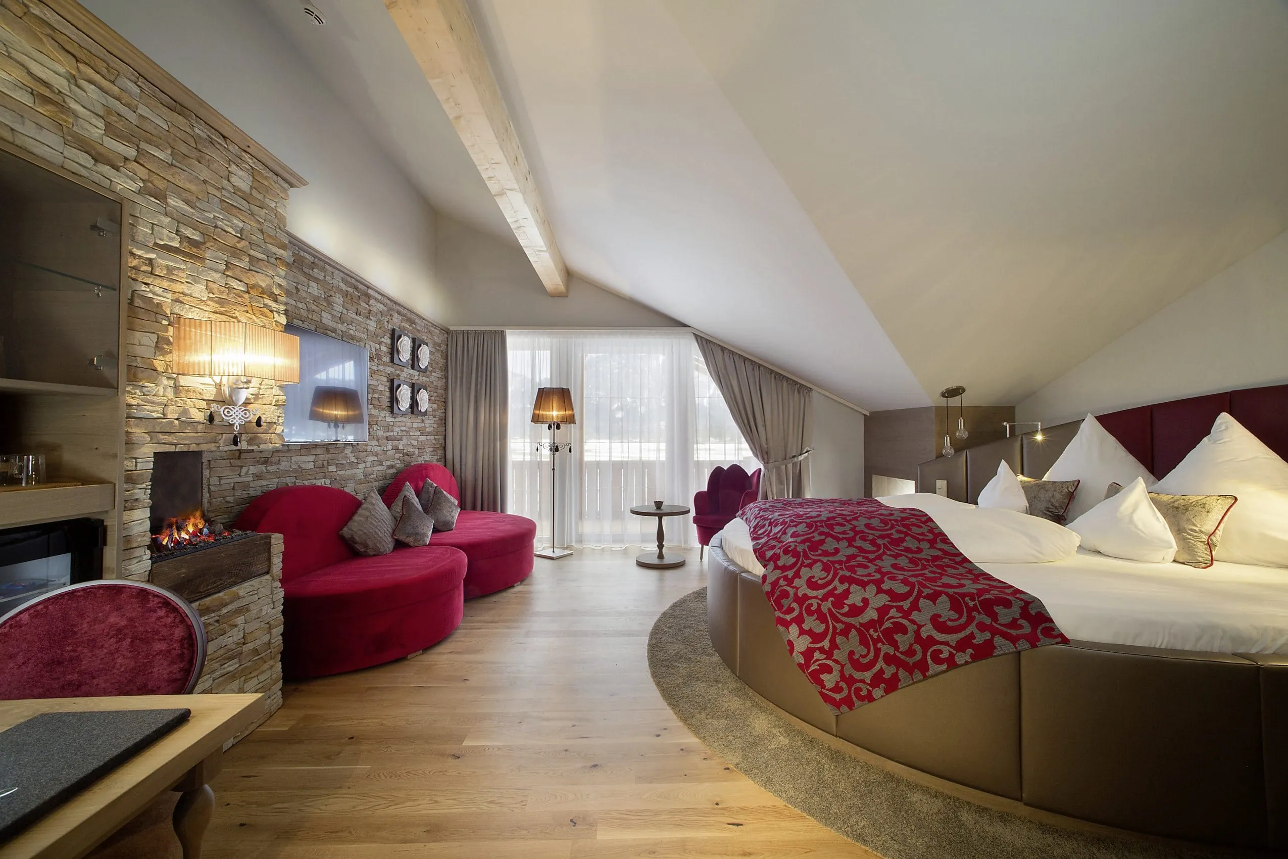 Suite mit heller Natursteinzierwand auf einer Seite und einem gro&szlig;en runden Bett auf der anderen Seite. Am Ende des Raums befindet sich ein Balkon.