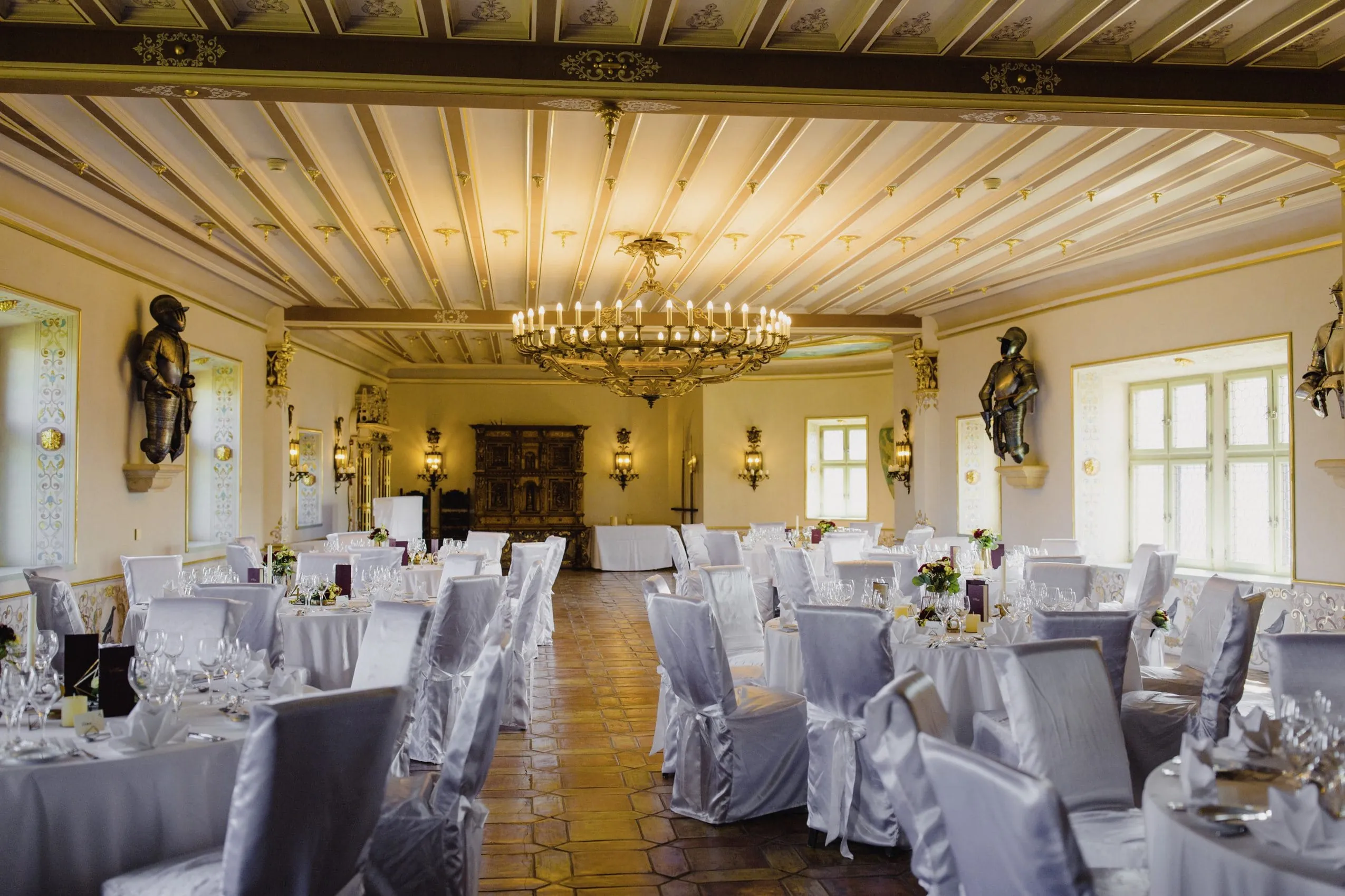 Der Renaissance-Saal mit gro&szlig;em Kronleuchter in der Mitte des Saals und Ritterr&uuml;stungen an den W&auml;nden. Im Saal stehen gro&szlig;e runde Tische mit wei&szlig;en Tischdecken und St&uuml;hle mit wei&szlig;en Hussen.