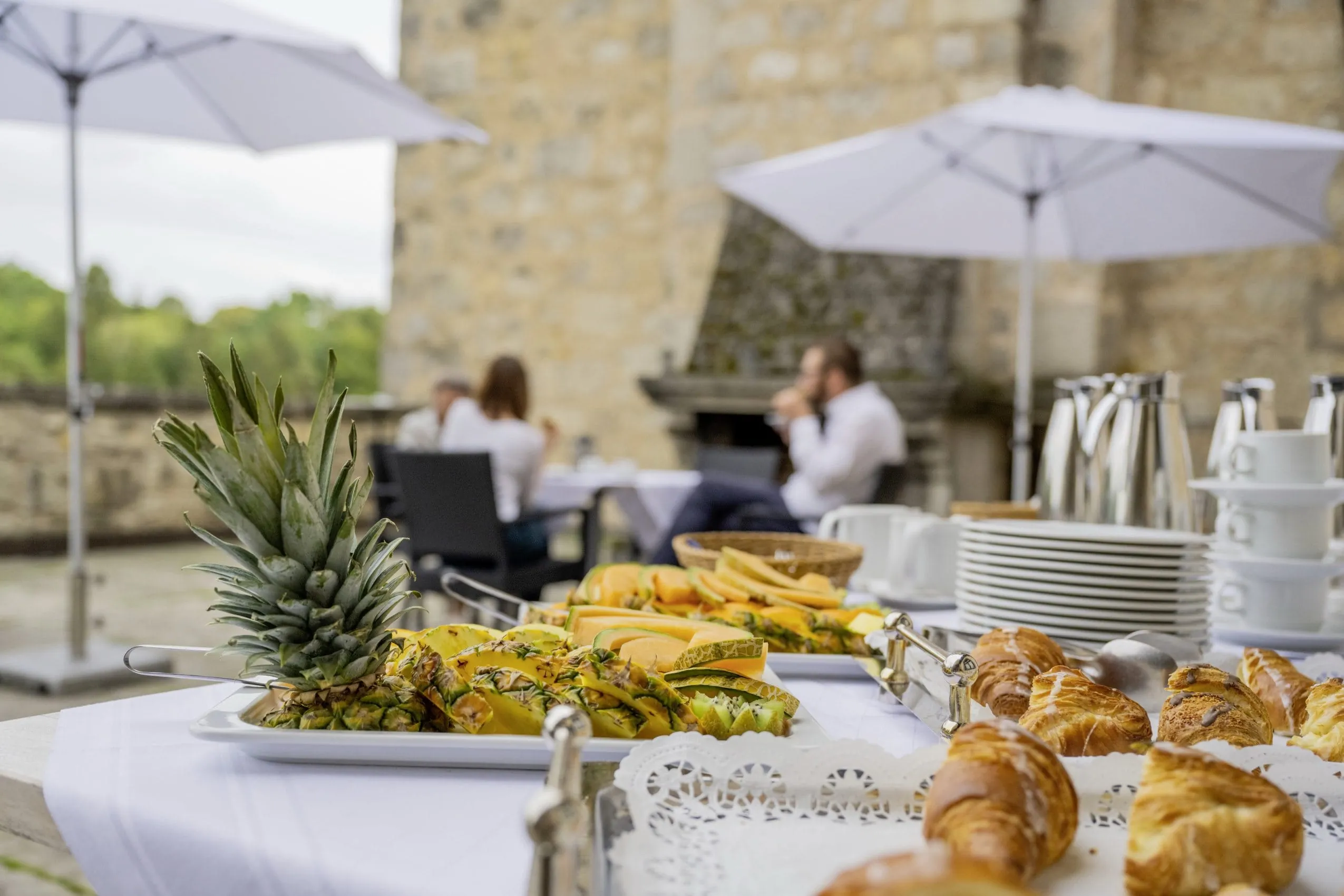Im Hintergrund unscharf die Burggem&auml;uer und Menschen beim Fr&uuml;hst&uuml;cken am Tisch. Im Vordergrund das Fr&uuml;hst&uuml;cksbuffet mit Croissants, Obstplatten, Kaffee, usw.