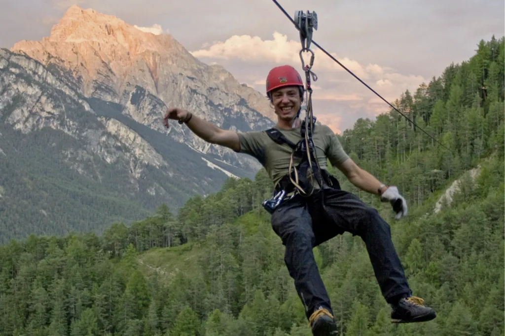 Ein junger Mann mit rotem Kletterhelm saust an einem dicken Stahlseil den Berg hinunter. Er schwebt dabei an Bergen und Wäldern vorbei.