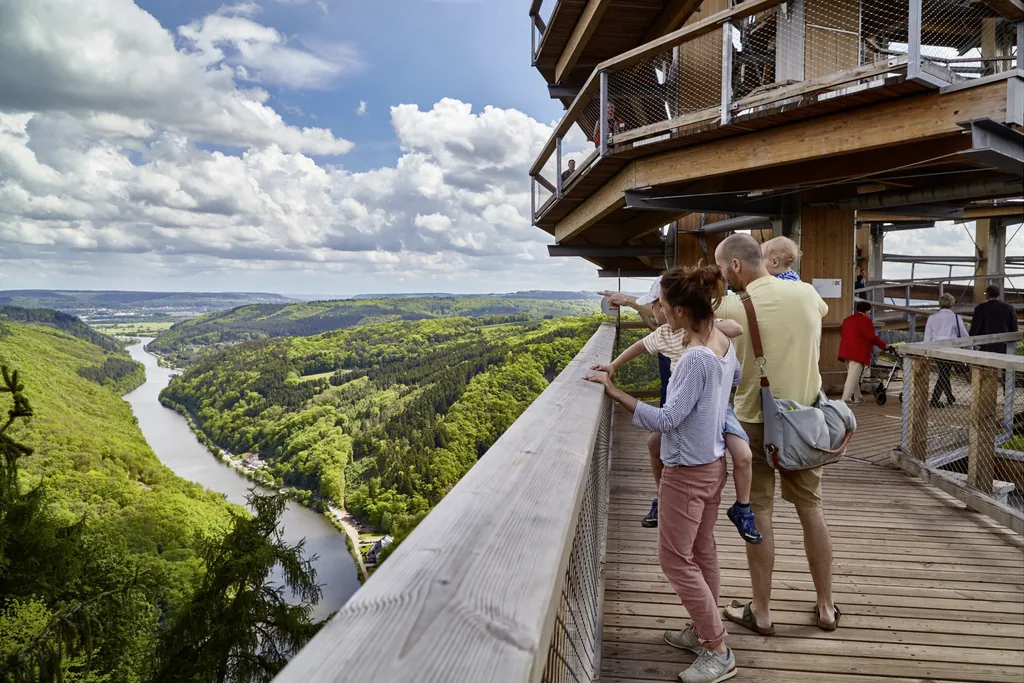 Eine Familie steht auf dem breiten Baumwipfelpfad aus Holz über den Bäumen mit Blick auf die umliegende Landschaft bestehend aus riesigen Wäldern und dem Fluss Saar, der sich durchschlängelt.
