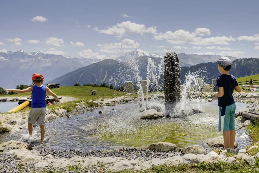 Zwei junge Buben in Tshirts und kurzen Hosen spielen an einem Teich mit Wasserspiel in der Mitte an dem Fontänen noch oben spritzen.