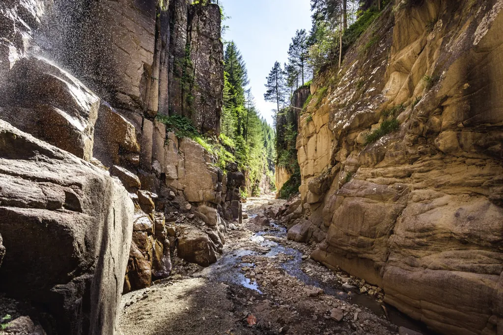 Riesengroße sandsteinfarbene Felswände. Zwischen ihnen bahnt sich ein Schotterweg sowie ein kleines Bächchen. Ganz oben an den Felswänden wachsen Bäume.